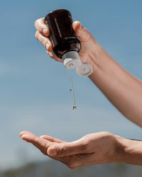 A person dispenses Rosie Jane's Calm Body + Bath Oil from a brown bottle onto their palm against a blue sky backdrop.