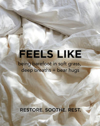 Rumpled white bed sheets with sunlight streaming in. Black text over the fabric reads: FEELS LIKE being barefoot in soft grass, deep breaths + bear hugs. RESTORE. SOOTHE. REST—with Rosie Jane Calm Body Milk hydrating body moisturizer.