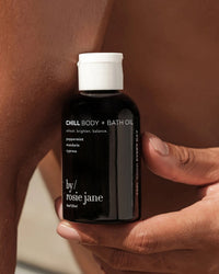 A person holds a bottle of Chill Body + Bath Oil by Rosie Jane, with peppermint, mandarin, and cypress, against their bare skin. The bottle has a black label with white text and a white cap.