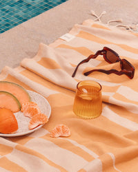 A patterned towel by the poolside is topped with sunglasses, a Saarde Kairos Medium Water Glass in Yellow Topaz, and a plate of sliced melon, orange, and tangerine.