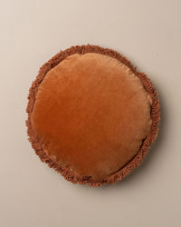 The Saarde Velvet Round Pillow w/ Feather Insert in Terracotta features a stylish fringe border and offers plush comfort, resting elegantly on a plain beige surface.
