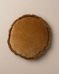 The Saarde Velvet Round Pillow w/ Feather Insert in Nutmeg features a vintage washed look, fringe edge, and plush feather down insert for comfort and timeless charm, shown styled against a simple light backdrop.