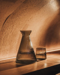 The Saarde Kairos Angular Carafe in Smoke, crafted from borosilicate glass, is filled with water and placed beside an empty tumbler on a wooden shelf under warm, ambient lighting.