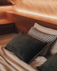 A neatly made bed with dark olive and striped pillows, including the Saarde Velvet Lumbar Pillow with Feather Insert in Dark Olive, rests against a wooden headboard softly lit by ambient lighting.