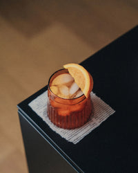 A glass of iced orange-colored drink, served in the Saarde Kairos Medium Water Glass in Dark Amber with an orange slice garnish, sits on a napkin atop a black table.