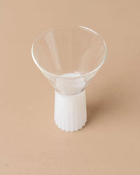 The Kairos Wine Glass in Opaque White by Saarde, crafted from clear borosilicate glass with a fluted white base inspired by Ancient Greek design, stands elegantly on a beige surface.