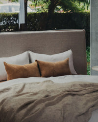 A neatly made bed with white and brown pillows, a beige blanket, the Saarde Velvet Lumbar Pillow w/ Feather Insert in Nutmeg, and a large window revealing greenery outside.