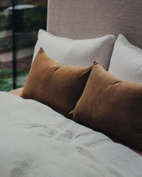 A neatly made bed with white bedding features two white pillows and Saarde Velvet Lumbar Pillows with feather inserts in Nutmeg, while a window and greenery provide a serene backdrop.