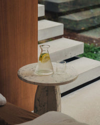 The Saarde Kairos Angular Carafe in Clear, filled with lemon water and paired with a tumbler, sits on a round marble side table next to steps leading outside.