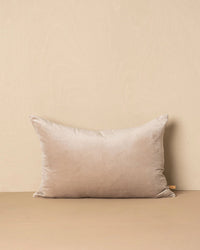 The Saarde Velvet Lumbar Pillow w/ Feather Insert in Stone stands upright against a matching wall and surface, featuring a beige velvet finish and a small fabric tag on the right side.