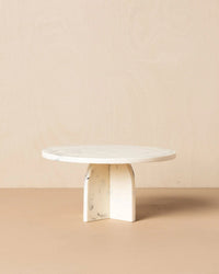 The Saarde Flow Resin Cake Stand in Merle features a round, white marble-look top with subtle grey veining and a thick X-shaped base, shown against a neutral beige background.