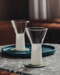 Two Saarde Kairos Wine Glasses in Opaque White, partially filled, rest on green coasters—one sits on a terrazzo table and the other is placed on a blue tray in the background.