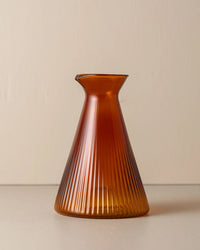 The Kairos Angular Carafe in Amber by Saarde, crafted from borosilicate glass with a flared base and narrow neck, is displayed on a plain surface against a beige background.