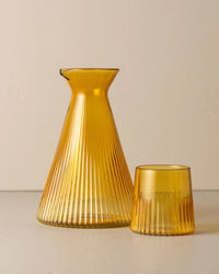 The Kairos Angular Carafe in Yellow Topaz by Saarde, featuring ribbed borosilicate glass with a narrow neck and flared base, stands beside a matching tumbler on a light surface against a neutral background.
