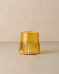 The Saarde Kairos Medium Water Glass in Yellow Topaz, featuring vertical grooves, sits on a light surface against a plain beige background.