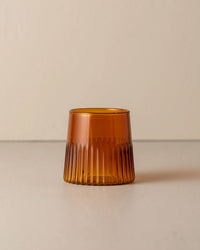 The Saarde Kairos Medium Water Glass in Dark Amber, featuring vertical ridges and a wide shape, sits on a neutral surface with a plain background.