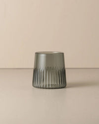 The Saarde Kairos Medium Water Glass in Smoke, featuring a smoky gray tint, is displayed on a beige surface with a matching background.