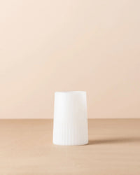 The Saarde Kairos Large Water Glass in Opaque White, featuring a cylindrical shape with a ridged lower half, stands upright on a beige surface against a light beige background.