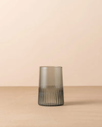 The Saarde Kairos Large Water Glass in Smoke, featuring a textured ribbed base, sits on a plain beige surface against a neutral background.