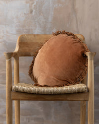 A wooden chair with a woven seat displays the Saarde Velvet Round Pillow with Feather Insert in Terracotta, adding warmth and texture against a neutral textured wall.