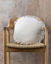 A Saarde Velvet Round Pillow w/ Feather Insert in Clay, featuring fringe edging, sits on a wooden chair with a woven seat against a textured beige wall.