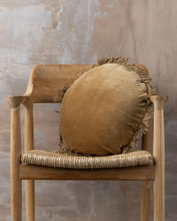 A Saarde Velvet Round Pillow with Feather Insert in Nutmeg rests on a wooden chair with a woven seat, set against a textured beige wall.
