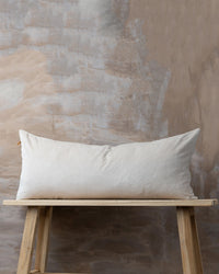 A Saarde Velvet Body Pillow with feather insert in clay rests on a wooden bench in front of a textured beige and gray wall.