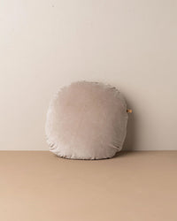 The Saarde Linen/Velvet Round Pillow with a feather insert in Stone is set against a plain beige wall and surface, featuring a small fabric tag.