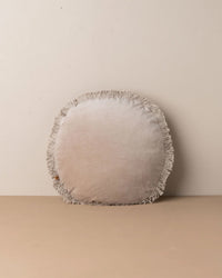 The Saarde Velvet Round Pillow with Feather Insert in Stone features fringe trim and vintage wash, shown upright against a light wall on a matching surface.