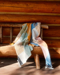 The Suave Campo - Blanket Roll by caminito, featuring a colorful patchwork of blue, yellow, pink, and white, is draped over a rustic wooden bench beside a log cabin wall with sunlight streaming in.