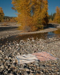 A caminito Suave Miel - Blanket Roll, handwoven in Mexico, rests on smooth river rocks beside a shallow stream, framed by tall trees with golden autumn leaves beneath a clear blue sky.