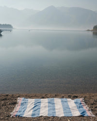 The caminito Suave Aire - Blanket Roll in blue and white stripes rests on a pebbled lakeshore, with misty mountains and trees visible across the calm water.