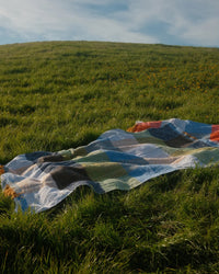 The Suave Kiwi - Blanket Roll by caminito lies spread out on tall green grass under a partly cloudy sky, atop a gently sloping hill adorned with yellow wildflowers in the background.