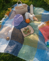 The caminito Suave Kiwi - Blanket Roll, handwoven in Mexico, is spread on grass with a wicker bag, purple cap, thermos, bread in paper, orange and kiwi—sunlight brightens this inviting indoor/outdoor scene.