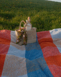 The caminito Suave Paseo - Blanket Roll, featuring red, white, and blue checks, is spread out on sunlit green grass with a picnic basket holding a floral scarf and baguette.