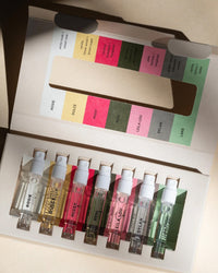 The Rosie Jane Fragrance Discovery Set includes seven labeled vials of clean perfumes arranged in a white box, with a chart matching each scent—Rosie, Dulce, Nissos, Remi, Bella Lou, Dylan, Lake—to its specific color.
