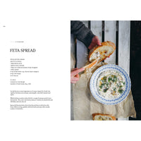 Using a knife, a person spreads creamy feta dip onto bread. Beside a rustic loaf, the herb-garnished dip sits in a bowl. Inspired by Four Seasons of Cabin Cooking from Cozy Publishing, this scene evokes simple cabin recipes and rustic charm.