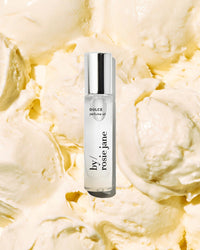 A clear glass roll-on Perfume Oil in Dulce by Rosie Jane, with a silver cap, is displayed against a creamy, textured backdrop reminiscent of vanilla ice cream.