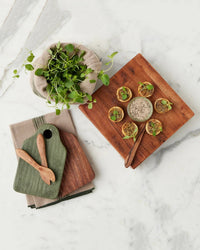 Overhead view of the etúHOME Dutch Small Cheese Board with Spreader in Spruce Green, arranged with pâté, herb-topped crackers, a wooden spreader, green microgreens, and two small boards with wooden spoons on marble.