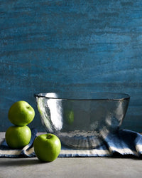 The Collective’s Pebbled Low Serving Bowl rests on a blue-striped cloth beside three green apples—two stacked, one apart—set against a textured blue backdrop.