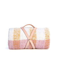 The caminito Suave Miel - Blanket Roll, a handwoven checkered throw in pink, white, and yellow from Mexico, is rolled up and fastened with a handmade leather strap against a white background.