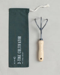 The Floral Society 3-Tine Cultivator, featuring a wooden handle, sits next to a dark green drawstring pouch labeled with its name—both shown on a light surface—ideal for garden tool enthusiasts.