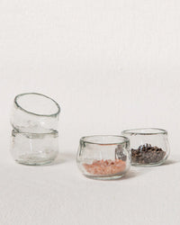 Four Pebbled Condiment Bowls by The Collective are arranged on a light surface; two are stacked empty, while the other two hold pink salt and peppercorns. Set of 4 recycled glass bowls.