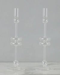 The etúHOME Clear Glass Candlesticks Set of 2 - Large features modern, geometric round and cylindrical shapes, displayed on a light surface with a gray background.