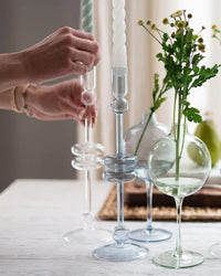 A person arranges the etúHOME Clear Glass Candlesticks Set of 2 - Large with white candles and a small glass vase of green foliage on a white table; each piece is handmade from recycled glass.