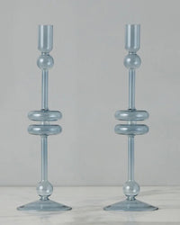 Two identical Denim Glass Candlesticks Set of 2 - Large by etúHOME, handmade from recycled glass with rounded, spherical accents, stand on a marble surface against a plain light background.