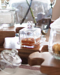 EtúHOME Bistro Glass Jam Jar, Square, by etúHOME, sits on a wooden serving board. Some jars are filled with jam, while a glass vase decorates the background.