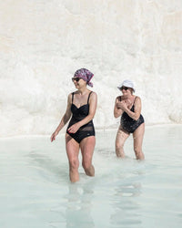 Two women enjoy Random House's Hot Springs, wading in shallow, light blue water with rocky scenery. One wears a patterned headscarf and sunglasses; the other dons a classic white hat as they splash and walk together.