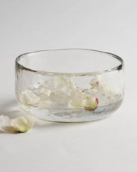 The Collective Pebbled Salad Bowl in clear recycled glass holds water with floating white flower petals, while a few petals and a bud rest on the white surface beside it.