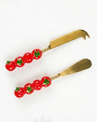 Two gold-toned Tomato Cheese Knives S/2 by Casa Amarosa, each featuring handles with three red ceramic tomatoes, are shown on a white background.
